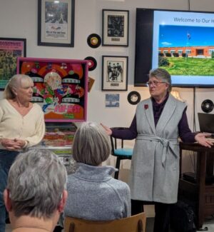 Linda & new Historical Society president Wilhelmina Women presenting with tv in the background to a small group of people sitting in chairs